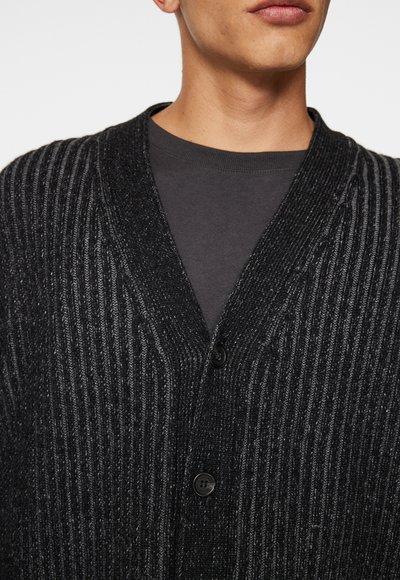 Sort strikket cardigan med vertikal ribbet tekstur, v-udskæring og to knapper. Bæres over en mørkegrå skjorte, minimalistisk design.