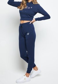 Navy modrý zkrácený svetr s logem na přední straně, doplněný o odpovídající jogger kalhoty s bočními kapsami a vyšitým logem. Bílé tenisky.