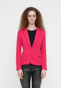 Γυναίκα με κατσαρά μαλλιά που φοράει ροζ σακάκι πάνω από μαύρο τοπ και σκουρόχρωμο υφασμάτινο παντελόνι, στέκεται μπροστά σε λευκό φόντο.