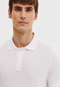 Biała koszulka polo z teksturowanej tkaniny, z kołnierzykiem i dwiema guzikami. Dyskretne logo na piersi, czysty i minimalistyczny design.