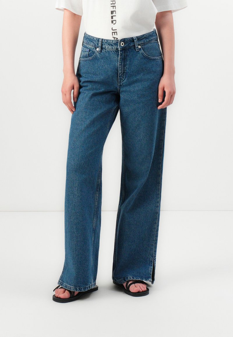 Ανοιχτό μπλε τζιν με φαρδιά πόδια από τζιν, με κλασικό σχέδιο πέντε τσεπών και σκισμένο στρίφωμα. Συνδυάζεται με λευκή μπλούζα και σανδάλια.