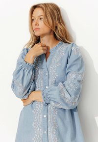 Chemise chambray bleu ciel ornée d'une broderie blanche et rose complexe sur le devant et les manches, avec des fermetures à boutons et des poignets froncés.