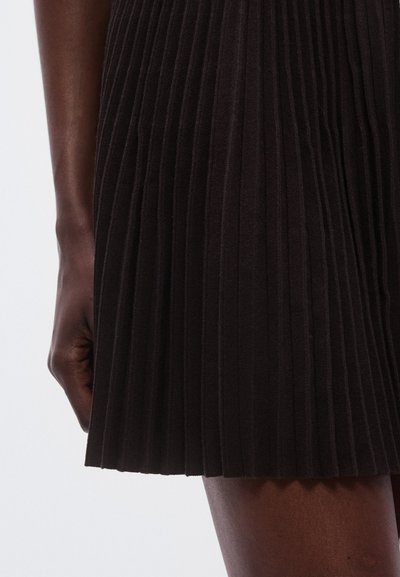 Jupe plissée noire en tissu texturé, avec des lignes verticales et un ourlet qui tombe au-dessus du genou, montrant une main tenant le tissu.