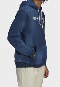 Námořnický modrý mikina vyrobená z měkké tkaniny, s přední kapsou, stahovacími šňůrkami a bílým logem na hrudi. Obsahuje žebrovaný lem.