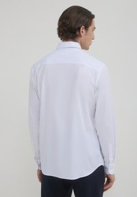 Camicia bianca a maniche lunghe con colletto classico, tessuto liscio e orlo curvo. Dotata di bottoni sui polsini e vestibilità sartoriale.
