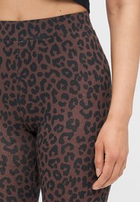Brahne tajice s crnim uzorkom leopard, glatke teksture i širokim pojasom, djelomično prikazane s rukom.