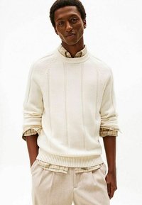 Maglione crema lavorato a maglia con orlo a coste e maniche allungate, sovrapposto a una camicia beige a quadri. Pantaloni chiari completano l'outfit.