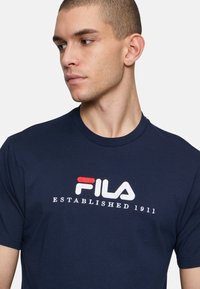 Camiseta de algodón azul marino con un logo bordado de FILA en blanco y rojo y el texto "ESTABLECIDO EN 1911" en el pecho. Mango corto y cuello redondo.