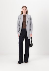 Sivi blejzer s jednim gumbom, smeđa majica i tamnoplave široke hlače. Dodaci uključuju malu crnu torbicu. Glatke teksture kroz cijeli outfit.