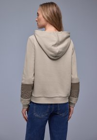 Femme portant un sweat à capuche beige avec imprimé léopard et patchs marron sur les manches, associée à un jean bleu, dos tourné sur un fond gris.