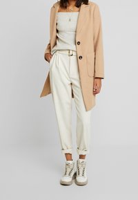 Beige Wollmischungsjacke mit schwarzen Knöpfen, hellgraues geripptes Off-Shoulder-Oberteil, cremefarbene tapered Hosen und weiße High-Top-Sneaker.