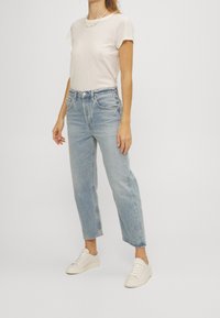 Ljusbeige t-shirt kombinerad med ljusblå raka jeans. Modellen har på sig vita sneakers. Bakgrunden är enkel och neutral.