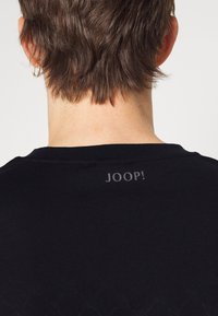 Černé bavlněné tričko s krátkým rukávem, s ribbovaným výstřihem a šedým potiskem loga "JOOP!" na zádech. Hladká textura.