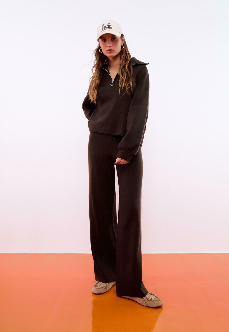 Maglione a maglia marrone con colletto a zip, abbinato a pantaloni a gamba larga coordinati; indossato con un cappellino beige con lettering nero e scarpe slip-on chiare.