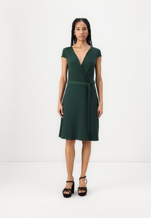 Robe verte avec un décolleté en V profond, des manches courtes et une taille nouée. Tissu lisse et léger, arrivant au genou, assortie à des sandales noires.