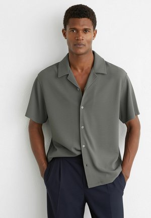 Młody mężczyzna w krótkim, szarym koszuli na guziki i ciemnych spodniach, stoi z rękami w kieszeniach na tle białej ściany.