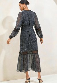 Musta pilkullinen midi-mekko, jossa on läpinäkyvää kangasta, pitkät hihat, kootut vyötärö ja kerroksellinen helma. Varustettu pyöreällä kaula-aukolla ja läpinäkyvillä yksityiskohdilla.