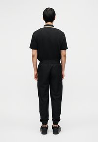 Μαύρο πόλο μπλουζάκι με κοντά μανίκια, συνοδευόμενο από μαύρο, υφασμάτινο jogger με ελαστική μέση και μανσέτες στα άκρα, φορεμένο με μαύρα παπούτσια.