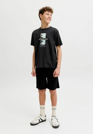 Teenagedreng med krøllet hår iført sort grafisk t-shirt, sorte shorts, cremefarvede sokker og hvide sneakers, der står på hvid baggrund.