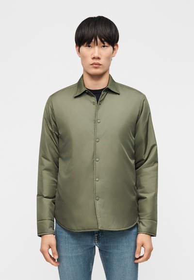 Jeune homme debout, face à la caméra, portant une veste matelassée olive verte boutonnée et un jean bleu sur un fond uni.