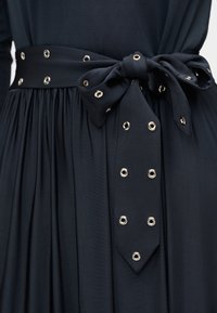 Μαύρο πλισέ φόρεμα με δεμένη υφασμάτινη ζώνη που διαθέτει ασημένια μεταλλικά κρικάκια τοποθετημένα σε σειρές γύρω από τη μέση.