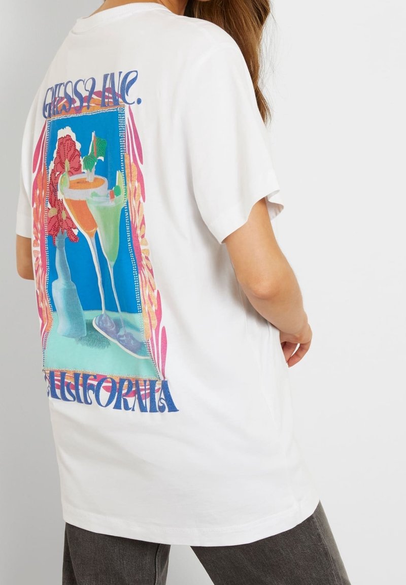 T-shirt blanc avec un graphisme coloré de cocktails, une bouteille et des fleurs, avec le texte "GUESS? INC." et "CALIFORNIA" dans le dos.