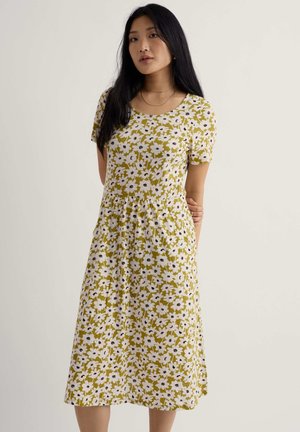 Młoda kobieta o długich czarnych włosach, ubrana w sukienkę z krótkim rękawem, sięgającą kolan, z białym kwiatowym wzorem na zielonym tle.