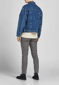 Giacca di jeans blu scuro indossata sopra una camicia a righe bianca e beige, abbinata a pantaloni grigi e scarpe nere, vista da dietro.