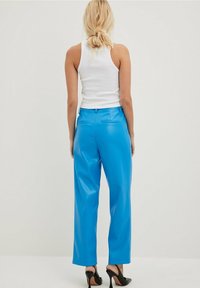 Blaue Kunstleder-Hose mit weitem Bein und glatter Textur. Verfügt über Gesäßtaschen und eine hohe Taille, kombiniert mit einem weißen Tanktop und schwarzen hohen Schuhen.