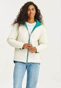 Prešívaná biela bunda s tyrkysovou kapucňou, na zips a s zelenými akcentmi. Modelka má v ruke kľúčenku, na sebe má bielu košeľu a džínsy.