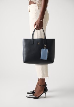 Kobieta w kremowych spodniach i czarnych szpilkach trzymająca czarną skórzaną torbę na ramię z dołączoną niebieską saszetką.