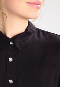 Chemise noire à col avec des boutons-pression argentés, présentant une texture lisse et des coutures visibles. Vue rapprochée de l'encolure et du tissu supérieur.