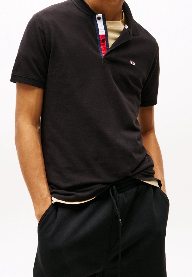 Polo noir avec un col agrémenté de touches rouges, blanches et bleues. Porté sur une chemise beige, associé à un pantalon noir. Coupe décontractée.