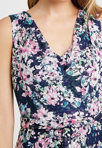 Blomsterklänning med marinblå bakgrund och rosa samt gröna blommor, med v-ringad design och ett tygband i midjan.