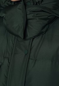 Mørkegrøn pufferjakke med en stor krave, trykknaplukninger, glat tekstur og synlige syninger på kroppen.