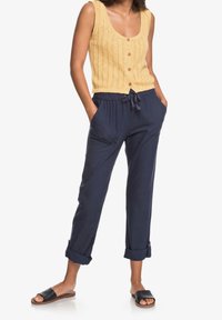 Débardeur jaune en tricot avec un motif texturé et des boutons en bois, associé à un pantalon bleu marine à coupe décontractée avec poches et revers. Sandales noires.