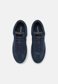 Marineblå sneakers med semsket skinn og syntetiske detaljer, rund tå, snøring foran, og hvit gummisåle. Merkevaren er synlig innvendig.