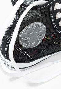 Baskets montantes en mesh noir avec lacets blancs, ornées d'un patch logo circulaire gris. Le design comprend des accents multicolores sur la surface.