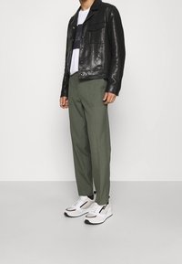 Černá kožená bunda s dvěma náprsními kapsami, kombinovaná s olivově zelenými kalhotami a bílými teniskami s černými akcenty a bočním zipem.