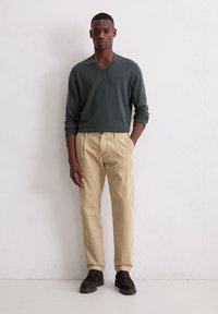 Maglione a V in verde scuro, abbinato a pantaloni in corduroy beige a pieghe. Presenta una vestibilità morbida e robusti scarponcini in suede neri.