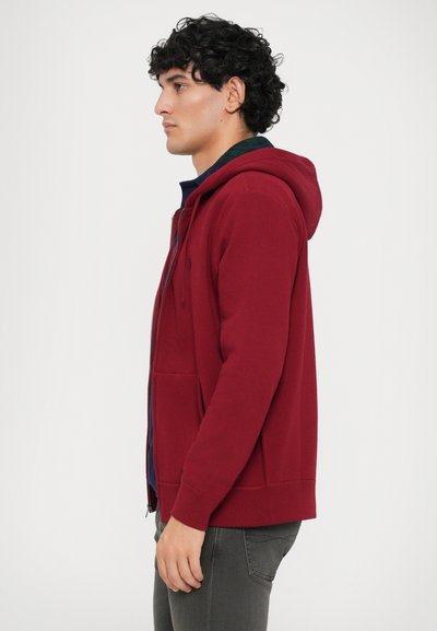 Hoodie vermelho com fecho à frente, bolso frontal, punhos canelados e capuz, usado sobre uma camisa azul. O tecido parece suave, com um ajuste casual.