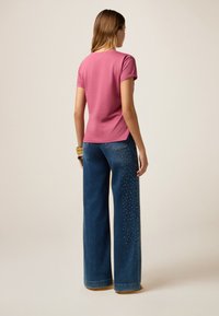 Camiseta rosa de manga corta con un escote redondeado, combinada con jeans de pierna ancha azules que tienen acentos de tachuelas en un lado.