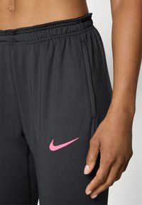 Sorte joggebukser med elastisk midje, med rosa Nike swoosh-logo på venstre side og sidelommer. Glatt tekstur.