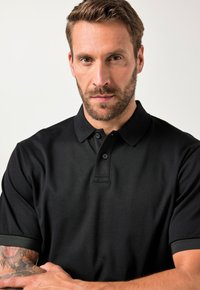 Hombre con cabello corto castaño y barba, vistiendo una camisa corta de manga corta negra con cuello, brazos cruzados, expresión neutral, fondo claro.
