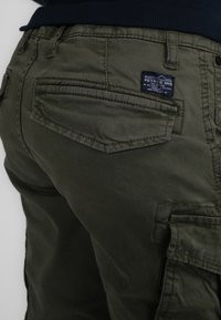 Olive green cargo pants with visible stitching, multiple pockets, and a fabric label positioned on the back pocket. Textured cotton material.