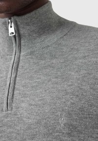 Maglione grigio con zip e colletto alto, caratterizzato da una texture a coste e un piccolo logo ricamato sul petto. Tessuto a maglia liscia.