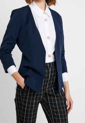 Kvinde iført navy blazer, hvid skjorte med guldknapper og sorte ternede bukser, hånd i lommen, mod en lys ensfarvet baggrund.