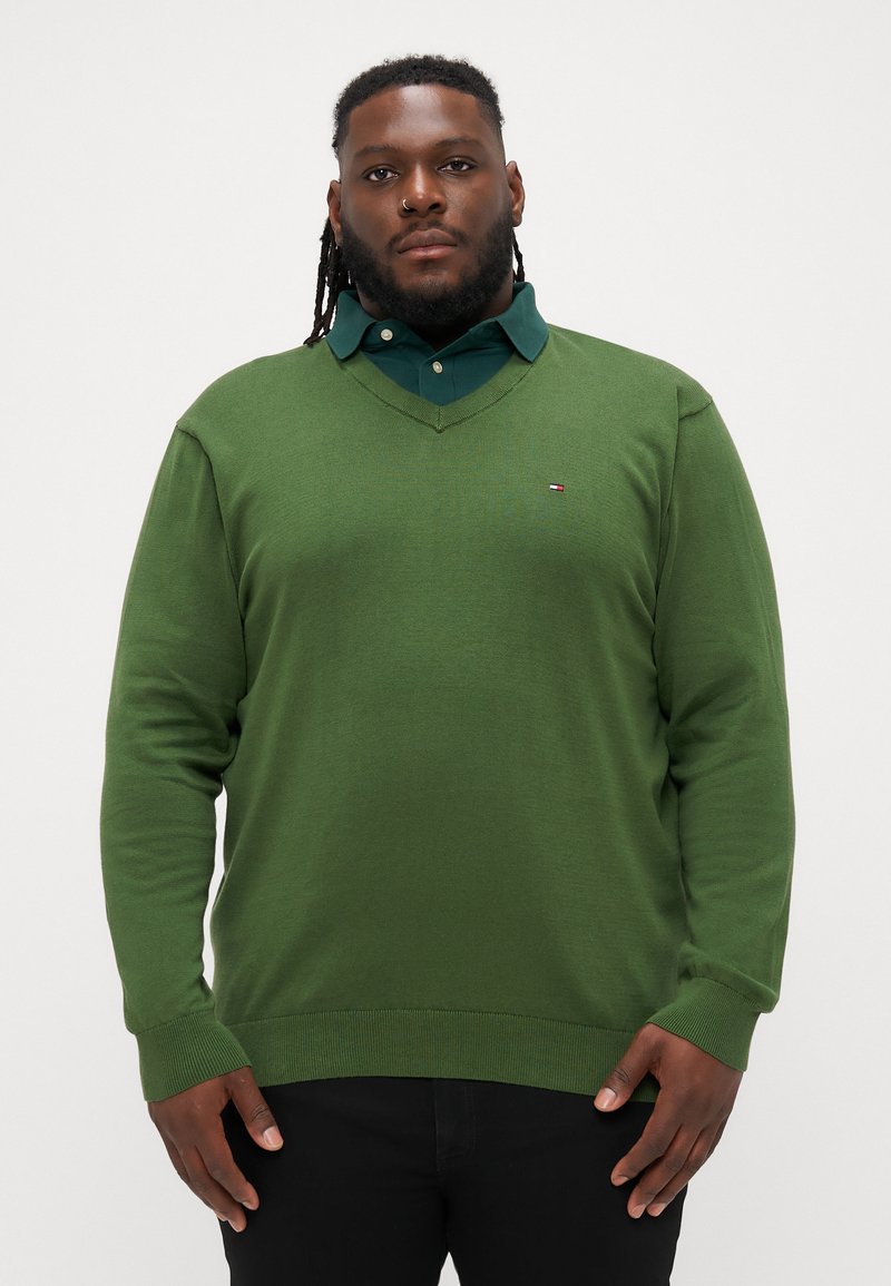 Zelený sveter s výstrihom do V vyrobený z pleteného materiálu, so zúženými manžetami a lemom. Pod ním je oblečená tmavozelená košeľa s golierom. Malé logo na hrudi.