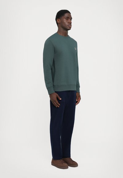 Sudadera verde oscura con cuello y puños acanalados, combinada con pantalones de color azul marino y zapatillas marrones. Detalle de logo sutil en el pecho.