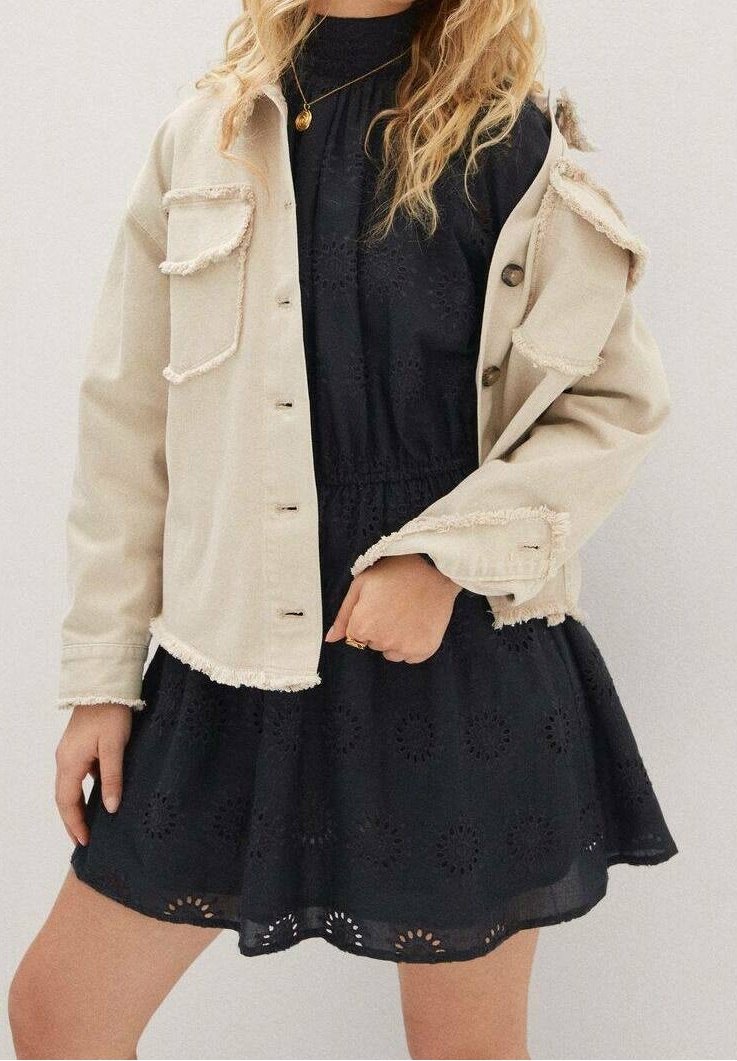 Beige denimjakke med taktile, frynsete kanter, fire lommer og knapper, lagd over en sort broderet kjole med cirkulære mønstre.